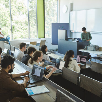 Menschengefüllter Hörsaal, während einer Vorlesung, mit einem Professor der vor einer großen Leinwand steht und eine Vorlesung hält.