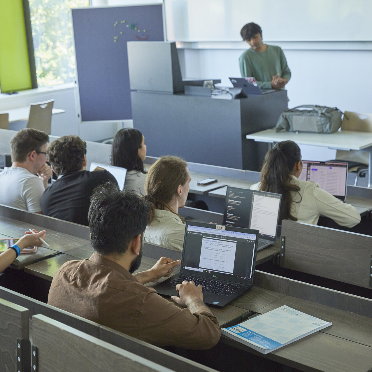 Menschengefüllter Hörsaal, während einer Vorlesung, mit einer lehrenden Person, die vor einer großen Leinwand steht.