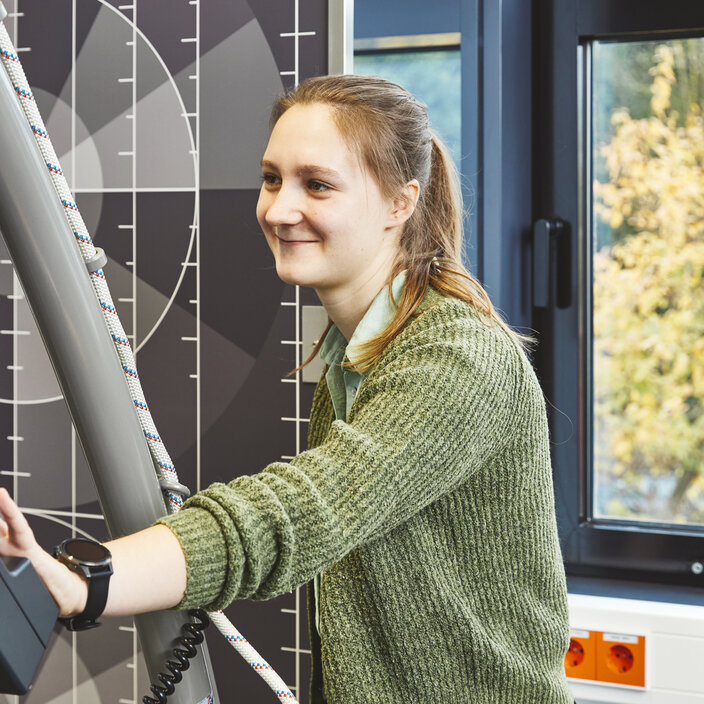 Eine lächelnde Studentin in einem grünen Oberteil steht an einem Kontrollpult und bedient ein großes Laufband. Links im Bild sieht man Teile des Laufbands und den Arm einer Person, die sich darauf bewegt.