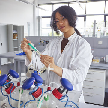 20260528_student_spritze_bioreaktor_01 Foto eines Studenten im weißen Kittel, der in einem Labor eine grüne Spritze an einem Versuchsaufbau mit Schläuchen sowie blauen und roten Verschlüssen bedient.