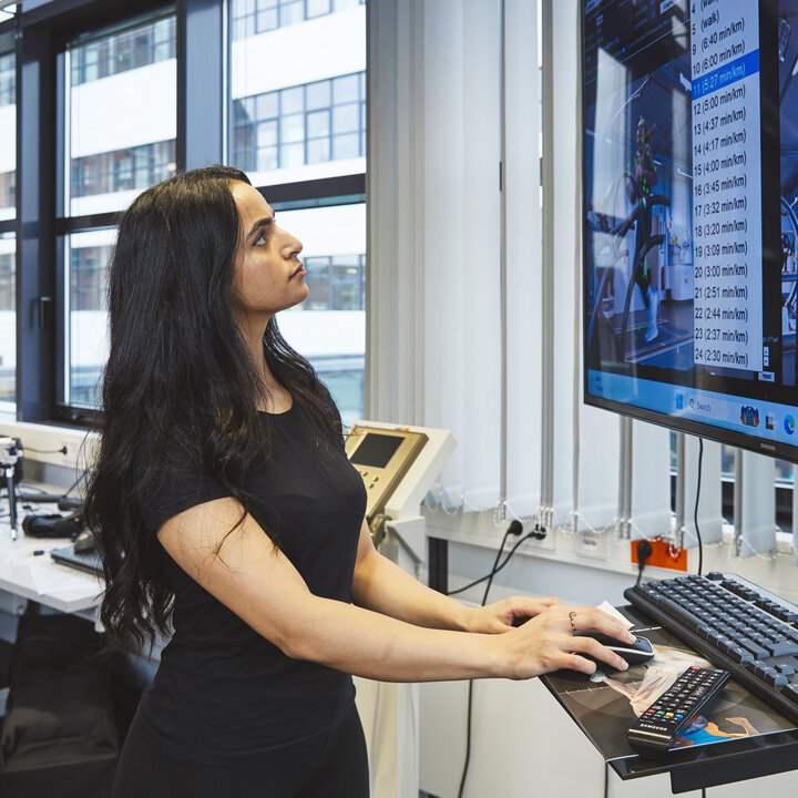 A young woman with long dark hair is standing at a standing desk in the laboratory. She is staring intently at a large monitor displaying detailed readings, video footage and a 3D animation of a person running. Her hand is resting on the computer mouse.