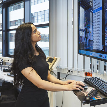 Eine junge Frau mit langen dunklen Haaren steht an einem Stehpult im Labor. Sie schaut hochkonzentriert auf einen großen Monitor, der detaillierte Messwerte, Videobilder und eine 3D-Animation einer laufenden Person anzeigt. Ihre Hand ruht auf der Computermaus.
