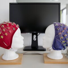 Two white model heads, each fitted with an EEG cap (red and blue) and numerous electrodes, face each other; there is a screen in the background.