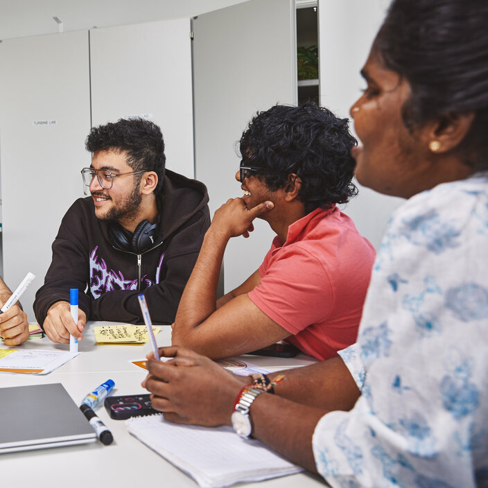 Four students are sitting at a table. Two of them are chatting and laughing together. The other two students are listening, smiling.
