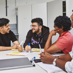 Vier Studierende sitzen an einem Tisch. Zwei von ihnen diskutieren miteinander und lachen sich an. Die anderen beiden STudierenden hören lächelnd zu.
