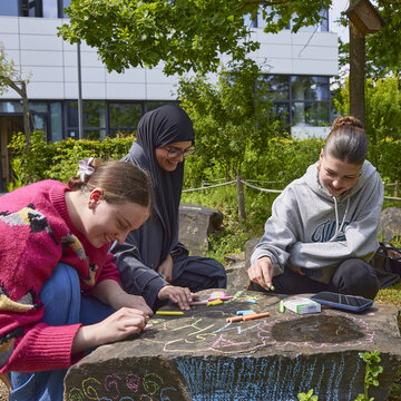 Drei Studierende sitzen in einem Garten, der sich auf dem Campus Kleve befindet und einer Außenanlage eines Kindergartens nachempfunden ist. Sie malen mit Kreide auf einen Stein und lachen dabei.
