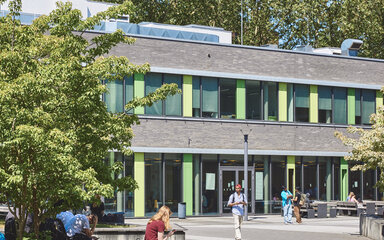Menschengefüllter Hochschulcampus, mit modernen Gebäuden und Grünflächen im freundlichen Tageslicht.