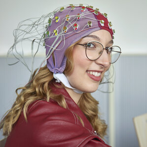 Studentin mit Brille trägt eine EEG-Kappe mit zahlreichen Elektroden und Kabeln.