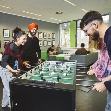 Vier Studierende stehen an einem Kicker-Tisch und spielen eine Partie. Im Hintergrund sind weitere Personen im Aufenthaltsraum zu sehen.