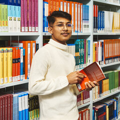Ein Student hält vor einem Bücherregal der Bibliothek Kleve ein Buch in den Händen und lächelt in die Kamera