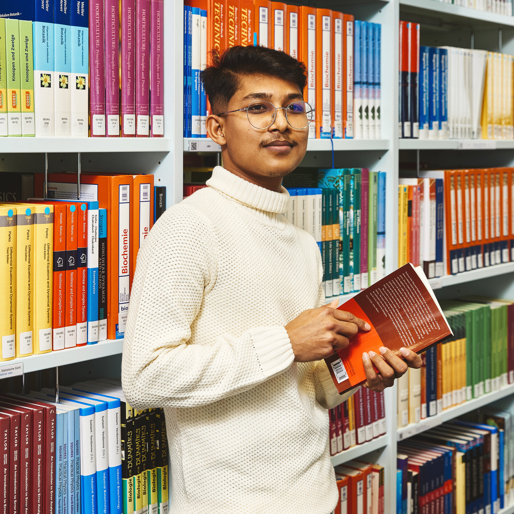 Ein Student hält vor einem Bücherregal der Bibliothek Kleve ein Buch in den Händen und lächelt in die Kamera