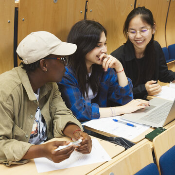 Drei Studierende sitzen im Hörsaal. Sie schauen auf den Laptop der Studentin, die in der Mitte sitzt. Sie sehen fröhlich aus.