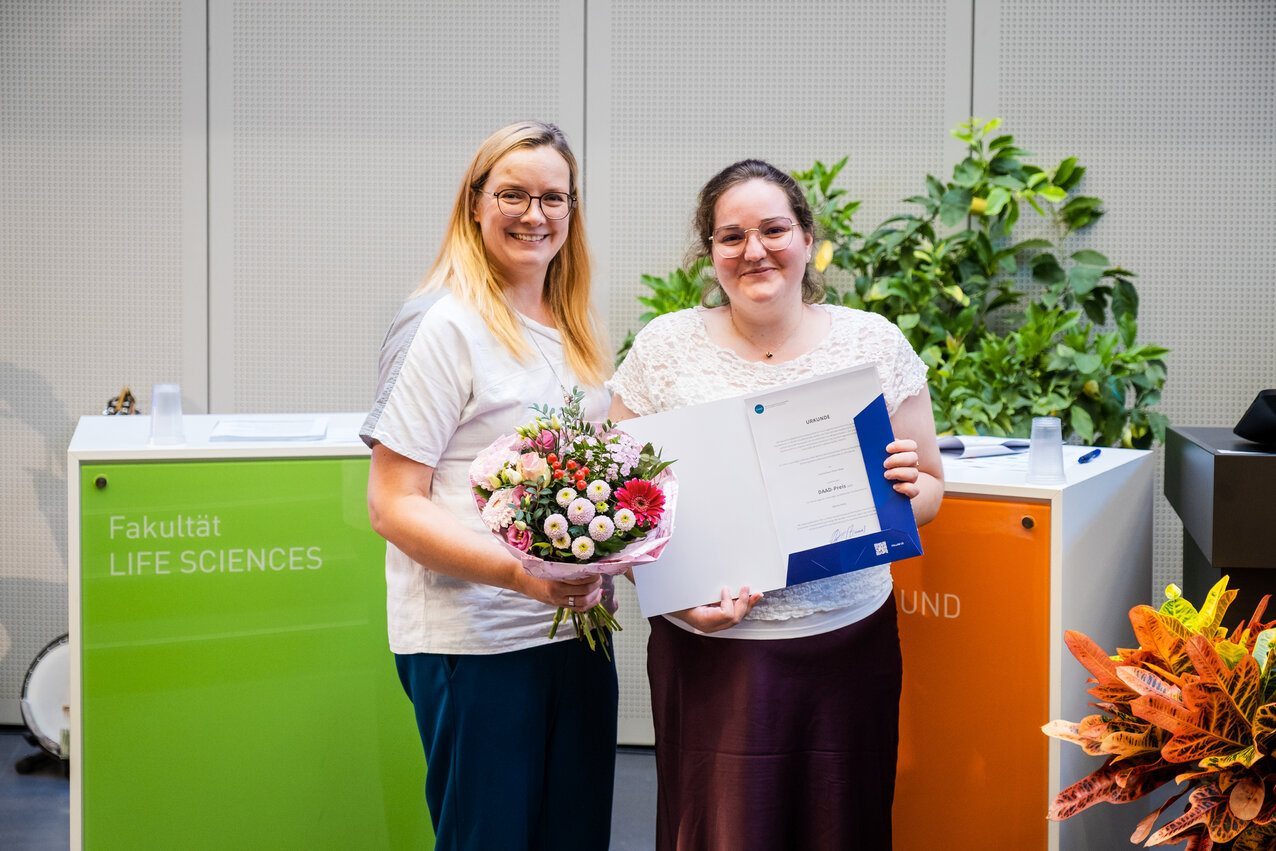 Zwei Frauen stehen vor dem Aufsteller der Fakultät Life Sciences, eine hält einen Blumenstrauß, die andere eine Urkunde.