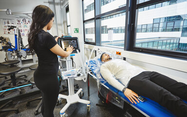 Calorimetric measurement in the laboratory – a student monitors a subject lying down and wearing a breathing hood A young man is lying relaxed on a lounger, wearing a transparent hood covering his head and upper body. A female student dressed in black is standing beside him, attentively operating the touchscreen of a medical monitoring device connected to the hood.