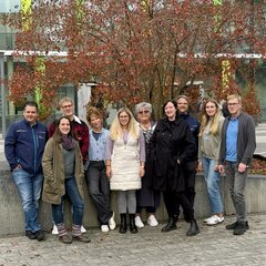 Vor einem herbstlich rot gefärbten Baum steht eine fröhliche Gruppe Personalratsmitglieder auf dem Campushof und lächelt in die Kamera. Im Hintergrund ist die Bibliothek des Campus Kamp-Lintfort mit ihren grünen Fensterrahmen zu sehen.