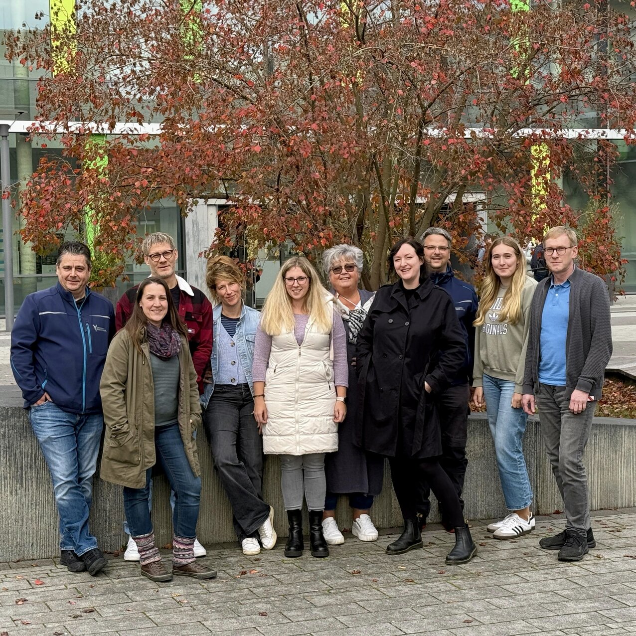 Vor einem herbstlich rot gefärbten Baum steht eine fröhliche Gruppe Personalratsmitglieder auf dem Campushof und lächelt in die Kamera. Im Hintergrund ist die Bibliothek des Campus Kamp-Lintfort mit ihren grünen Fensterrahmen zu sehen.