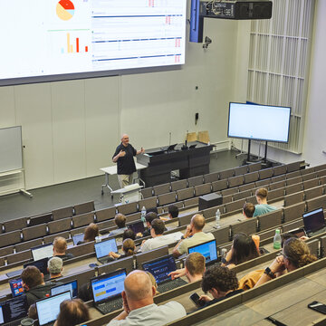 Menschengefüllter Hörsaal, während einer Vorlesung, mit einem Professor der vor einer großen Leinwand steht und eine Vorlesung hält.