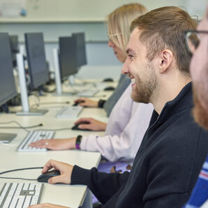 Nahaufnahme eines lächelnden Studenten in einer Reihe von Studierenden an Computerarbeitsplätzen.
