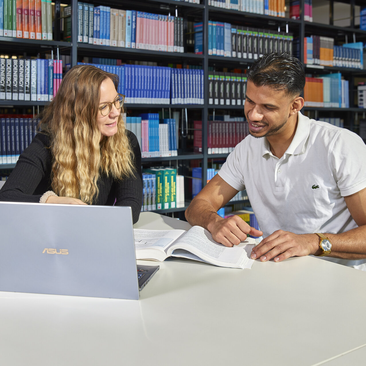 Two students are sitting together at a table in the library, working on a laptop and a book.