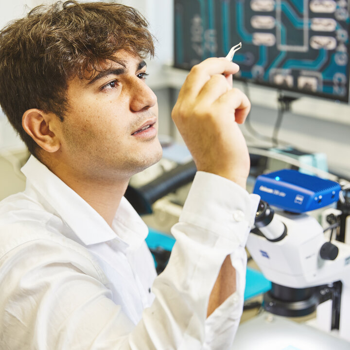 Ein Studierender im Laborkittel sitzt in einem Labor für Elektronik. In der Hand hält er eine Pinzette, mit der er ein kleines Bauteil betrachtet.