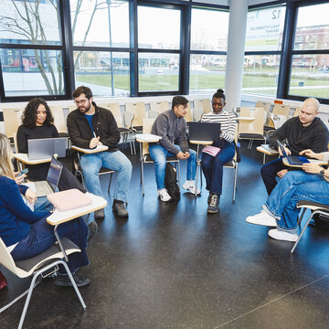 Stuhlkreis mit Laptops - Ein eigenes Unternehmen aufbauen Eine Gruppe von sechs Studierenden sitzt in einem Stuhlkreis in einem Raum mit Fensterfront. Sie haben Laptops auf den Schoßtischen und diskutieren Projektinhalte.