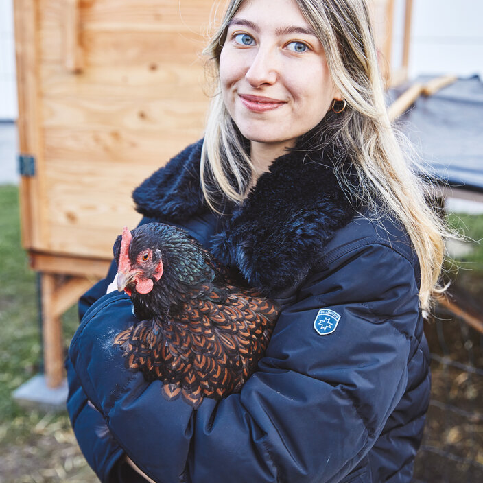 Studentin mit Huhn - Echter Praxisbezug im Studium Eine junge Studentin mit blonden Haaren steht draußen in einer Winterjacke und hält lächelnd ein braun-schwarz gefiedertes Huhn im Arm. Im Hintergrund ist ein hölzerner Hühnerstall zu sehen.