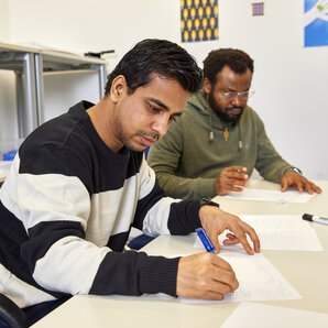Zwei Studierende arbeiten gemeinsam an einem Tisch mit Papier, Stiften und weiteren Arbeitsmaterialien.