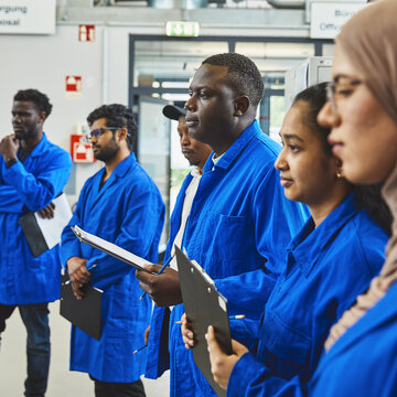 Sechs Studierende stehen im Labor und hören zu. Sie haben blaue Kittel an.