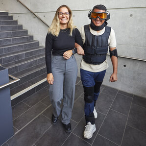 A student is wearing an ageing simulation suit and is being helped along by a female student.