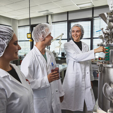 Studierende in Schutzkleidung an einer industriellen Mischanlage Drei Studierende in weißen Kitteln und Haarnetzen stehen an einer großen Edelstahl-Mischanlage. Ein Student hält ein Messgefäß mit roten Früchten, während eine Kommilitonin lächelnd an der Anlage hantiert.