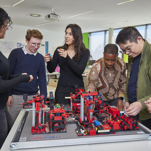 Sechs Studierende arbeiten gemeinsam an einer Modellfabrik mit verschiedenen technischen Modulen.