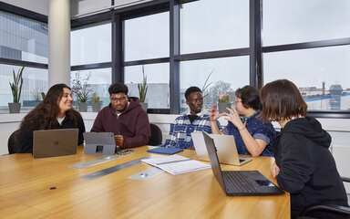 Mehrere Studierende sitzen um einen Tisch herum vor ihren Laptops miteinander ins Gespräch verteift