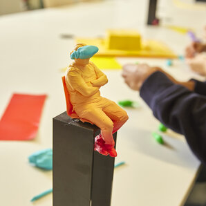 Nahaufnahme einer orangefarbenen Knetfigur mit verschränkten Armen, die auf einem dunklen Sockel sitzt und Blau über den Augen trägt.
