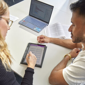 Two students are sitting at a table together, working on a tablet and a laptop.