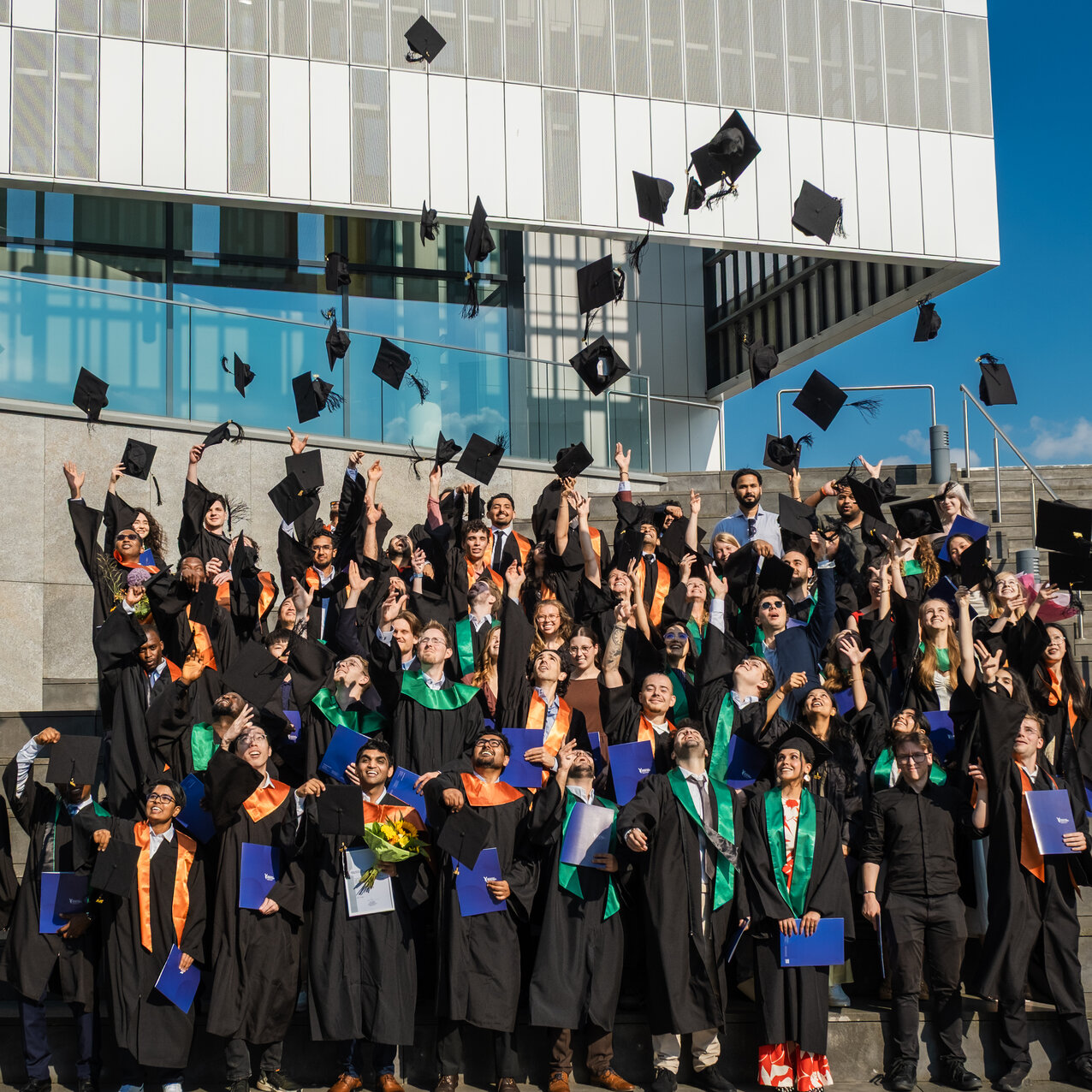 Around 70 graduates are standing on the steps in front of a building. They are wearing black gowns with orange and green sashes and are throwing their hats into the air. 
