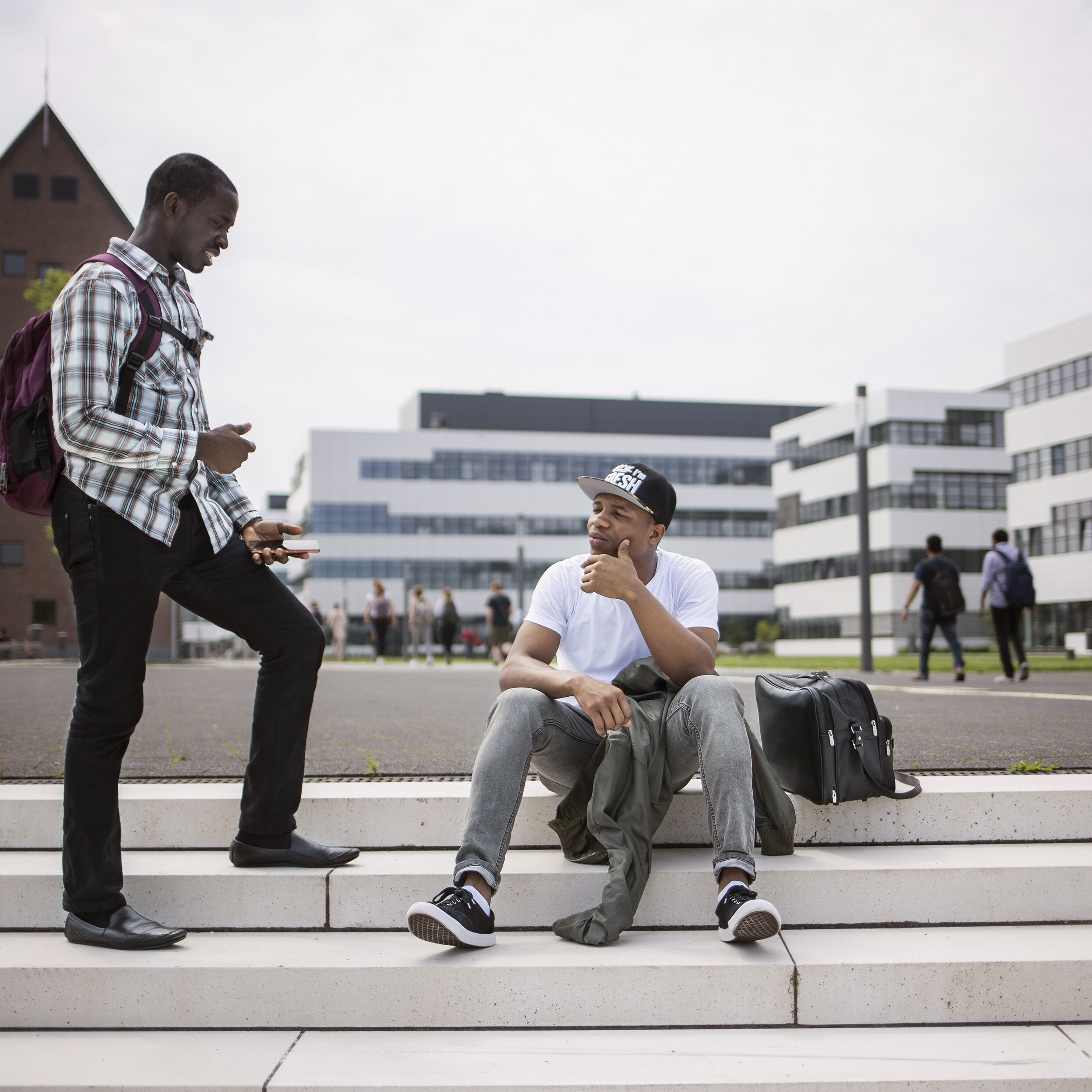 2 Studenten befinden sich auf dem Campus und unterhalten sich, im Hintergrund sind die Campusgebäude zu sehen