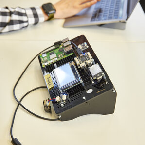 Nahaufnahme eines elektronischen Prototyps auf einem Tisch, im Hintergrund tippt eine Person auf einem Laptop.