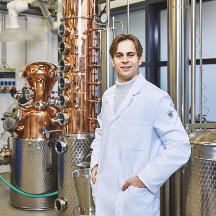Student vor industrieller Laboranlage - Ein lächelnder Student im Laborkittel steht vor einer großen Destillationsanlage aus Kupfer und Edelstahl Ein junger Student im weißen Laborkittel steht lächelnd mit den Händen in den Kitteltaschen in einem Labor. Direkt hinter ihm ragt eine große, komplexe Industrieanlage aus glänzendem Kupfer und Edelstahl auf.