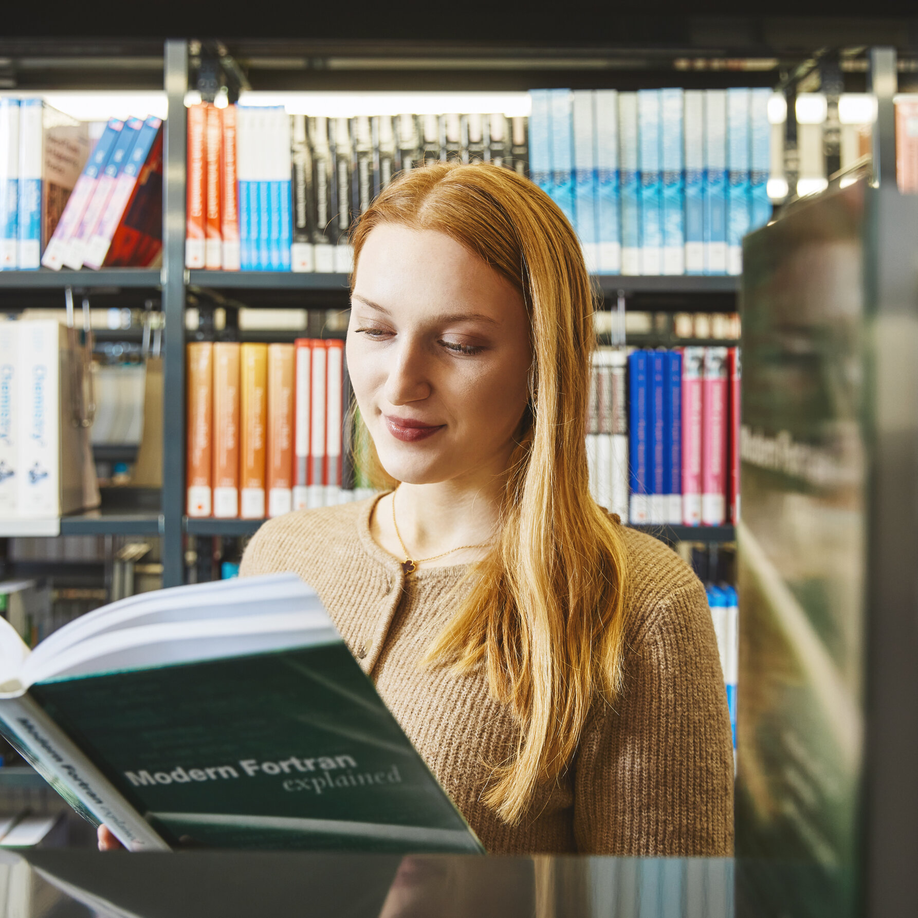 A student is reading a book and is pictured standing in front of a bookshelf in the library in Kamp-Lintfort.