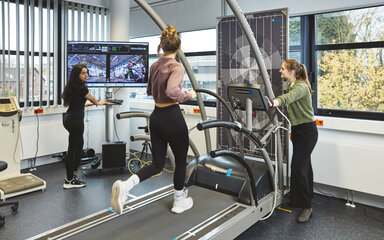 Eine Studentin in schwarzer Sportkleidung steht vor einem großen Monitor und bedient Maus und Tastatur. Auf dem Bildschirm sind Kamerabilder und Messdaten einer Frau zu sehen, die auf einem Laufband im selben Raum joggt.