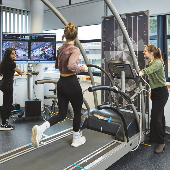 Eine Studentin in schwarzer Sportkleidung steht vor einem großen Monitor und bedient Maus und Tastatur. Auf dem Bildschirm sind Kamerabilder und Messdaten einer Frau zu sehen, die auf einem Laufband im selben Raum joggt.