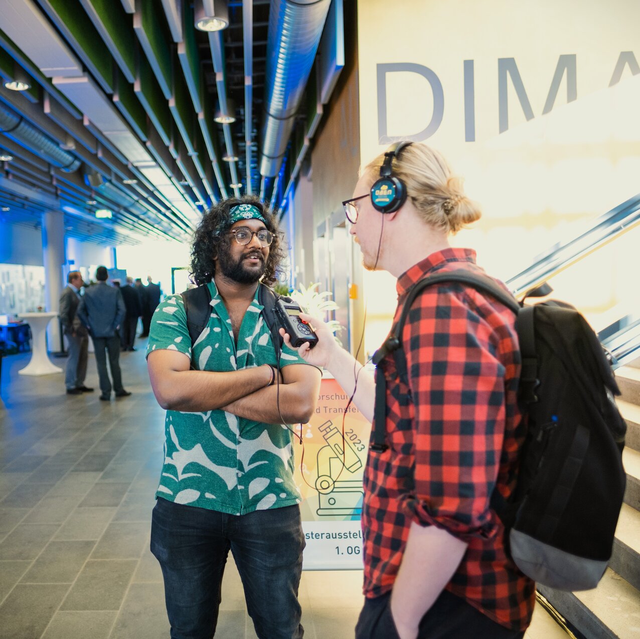 Zwei Personen stehen im Foyer des Audimax vor einer Treppe. Eine Person trägt Kopfhörer und hält der anderen ein Aufnahmegerät entgegen. 