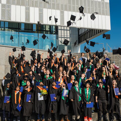 Graduates in academic gowns toss their hats into the air on the steps in front of the Audimax.