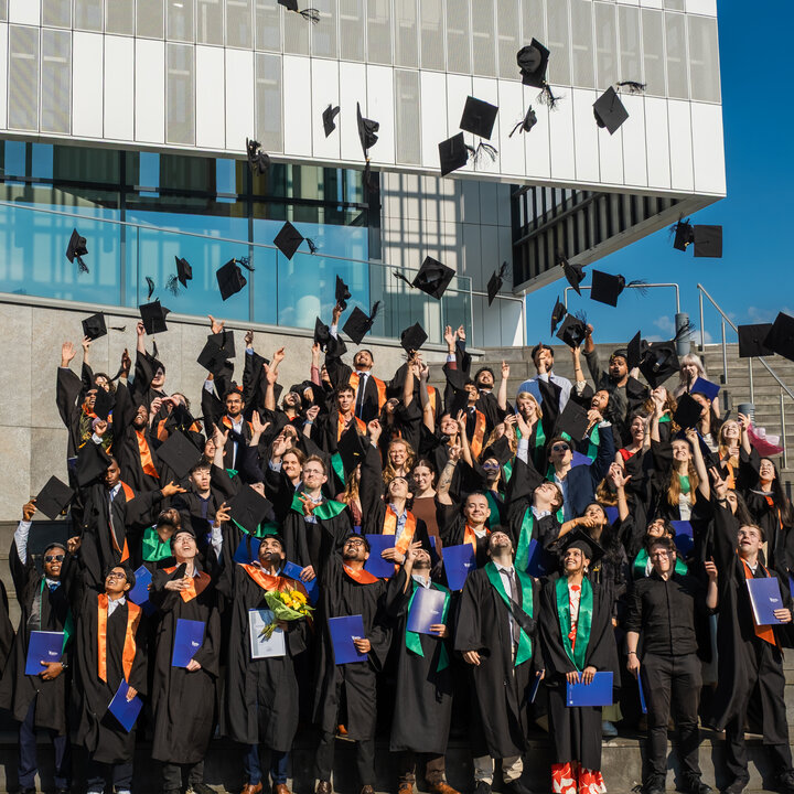 Graduates in academic gowns toss their hats into the air on the steps in front of the Audimax.