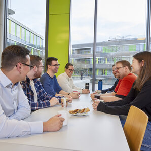 Studierende sitzen an einem Tisch in einer Mensa, halten Tassen und unterhalten sich.