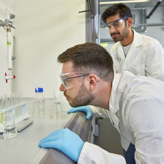 Zwei Studierende stehen im Chemielabor, sie tragen weiße Laborkittel und blaue Latexhandschuhe. Ein Studierender betrachtet aus der Nähe eine Reihe von Reagenzgläsern in einem Holzständer. Der andere Studierende steht hinter ihm und schaut ebenfalls auf die Reagenzgläser, in denen verschiedenfarbige Flüssigkeiten sind.
