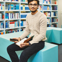 Ein Student sitzt mit Handy und Buch in der Bibliothek Kleve und lächelt in die Kamera
