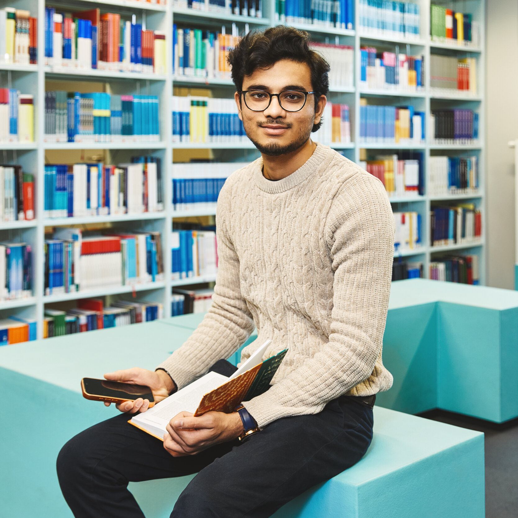 Ein Student sitzt mit Handy und Buch in der Bibliothek Kleve und lächelt in die Kamera