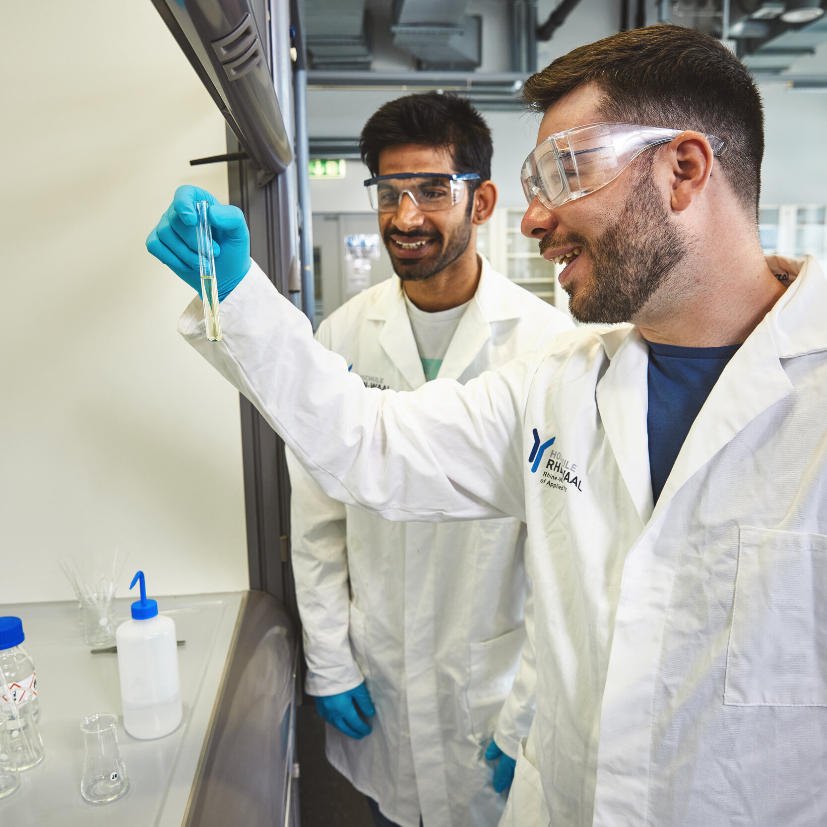 Zwei Studierende stehen in einem Chemielabor. Sie tragen weiße Laborkittel und blaue Latexhandschuhe. Ein Studierender hält ein Reagenzglas in der Hand.