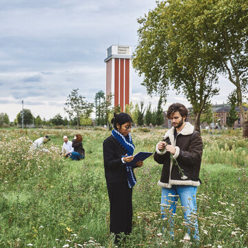 Zwei Studierende untersuchen Pflanzen auf dem Gelände der Landesgartenschau direkt neben dem Campus Kamp-Lintfort. Im Hintergrund sieht man den alten Förderturm der ehemaligen Zeche.